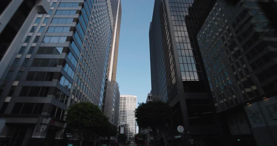 A drive through a city street lined with tall buildings some with reflective glass facades and others with concrete or brick under a bright blue sky with traffic lights and street signs visible as the perspective moves forward