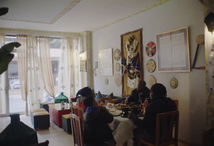 A family sit around a dining table in a restaurant with large windows decorative wall hangings and ornate ceiling trim as people walk outside
