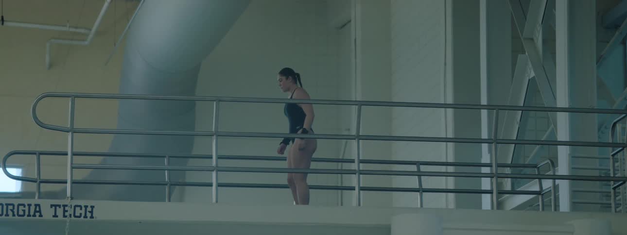 A female athlete swimmer in her swimsuit stands on a diving platform practicing her diving skills as she prepares to dive into the pool