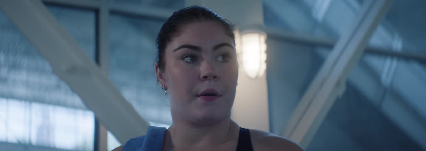 A young adult woman with her hair pulled back and a blue towel draped over her shoulder looks around with a focused expression in what appears to be a gym or athletic facility with large windows and structural beams015