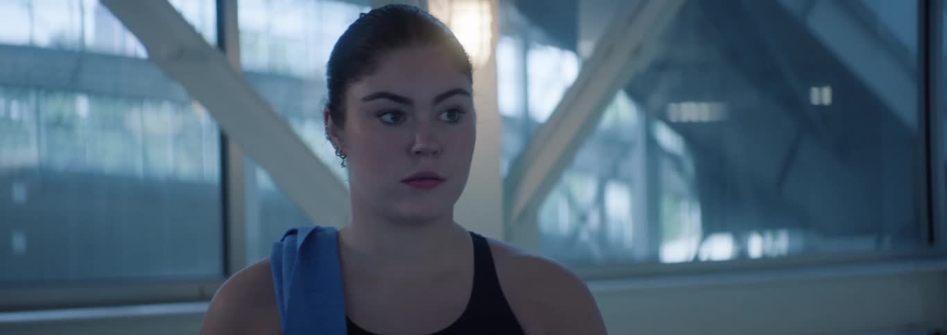 A young adult woman with her hair pulled back and a blue towel draped over her shoulder looks around with a focused expression in what appears to be a gym or athletic facility with large windows and structural beams