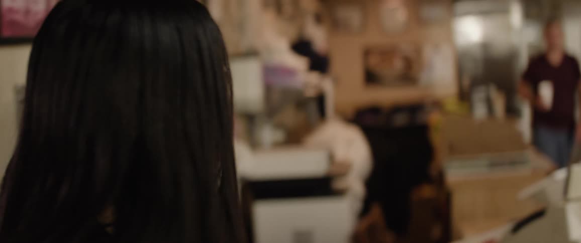 A young adult woman with long hair wearing a dark leather jacket and a light colored top walks into a casual diner or restaurant smiling as she passes through the entrance and moves into the dining area with other customers
