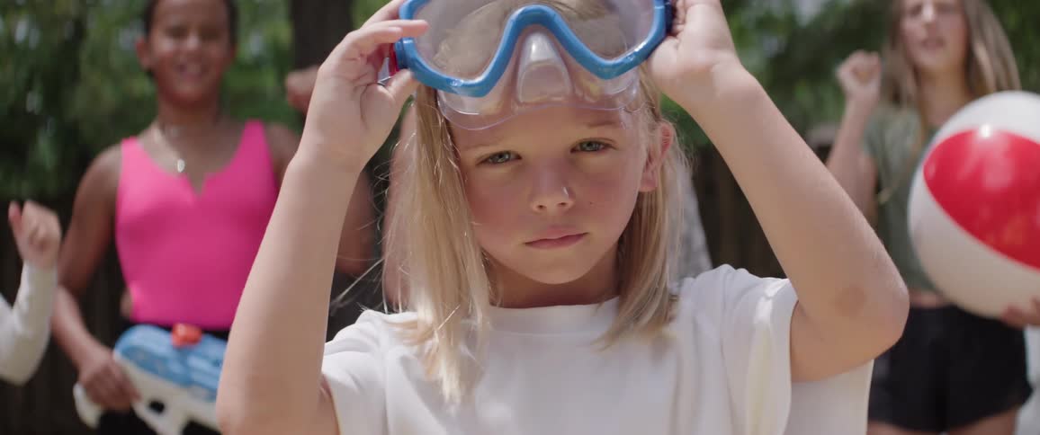 Little blonde girl taking off her swimming goggles with a concentrated expression and other people in the background cheering happily