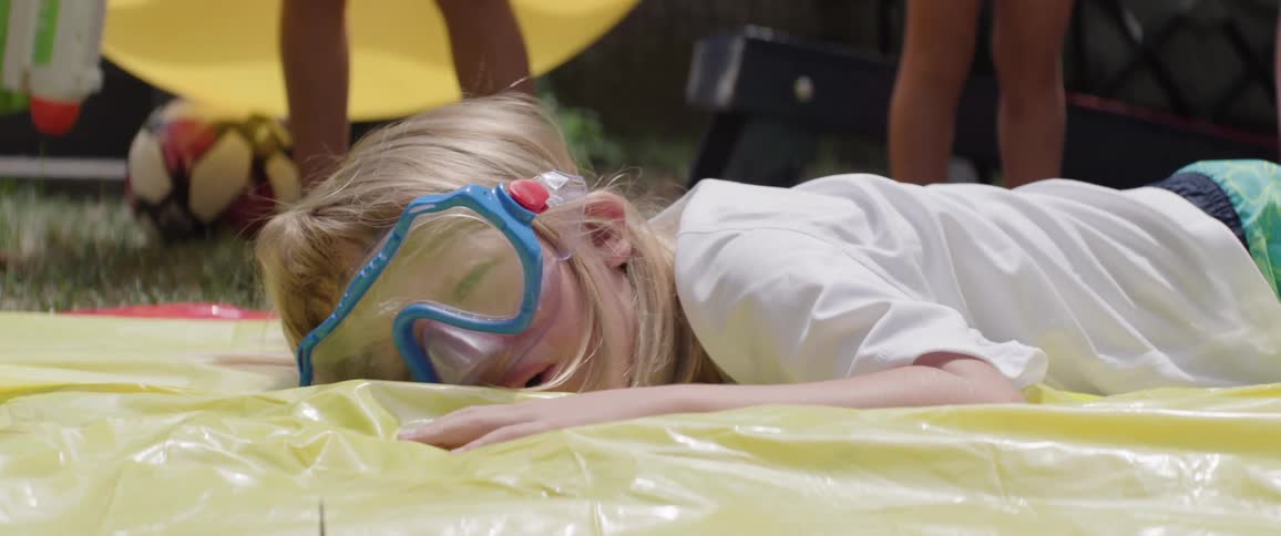 Blond child with swimming goggles on his face falling on an inflatable toy on the grass