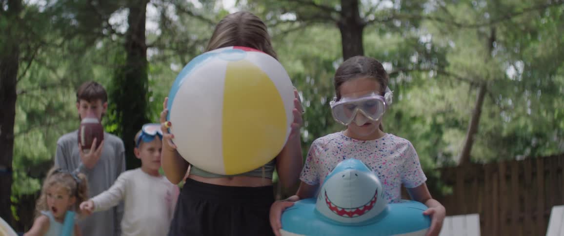 The video shows three children standing outdoors in a grassy area with two boys and a girl the girl on the right is wearing goggles and holding a blue inflatable shark