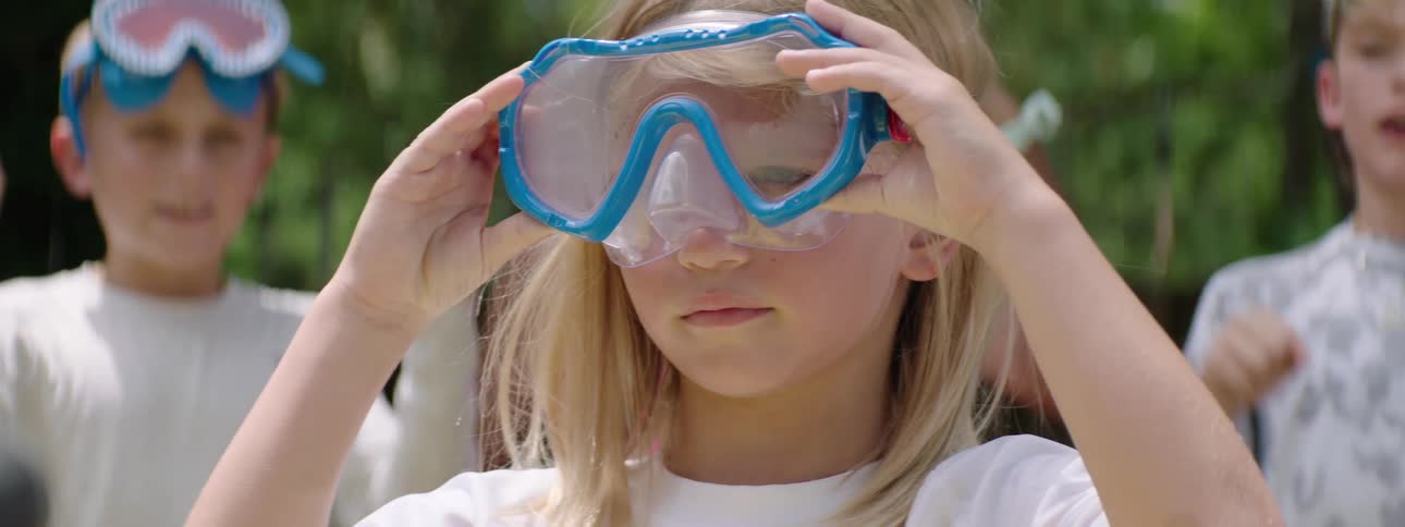 A young girl wearing swim goggles stands still with a thoughtful expression surrounded by other children blurred in the background