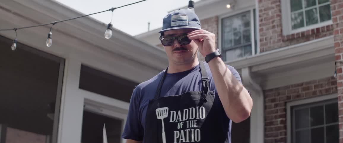Man in a bbq apron lowers his spatula and removes his sunglasses before making a comment