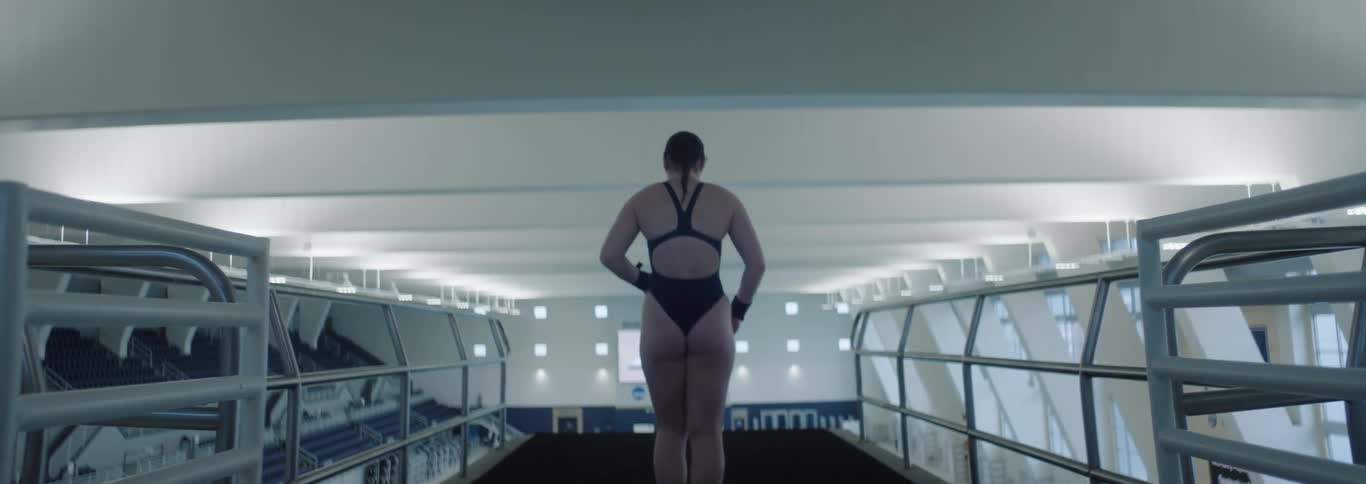 A woman in a black swimsuit with a towel over her shoulder walks toward an indoor swimming pool viewed from behind
