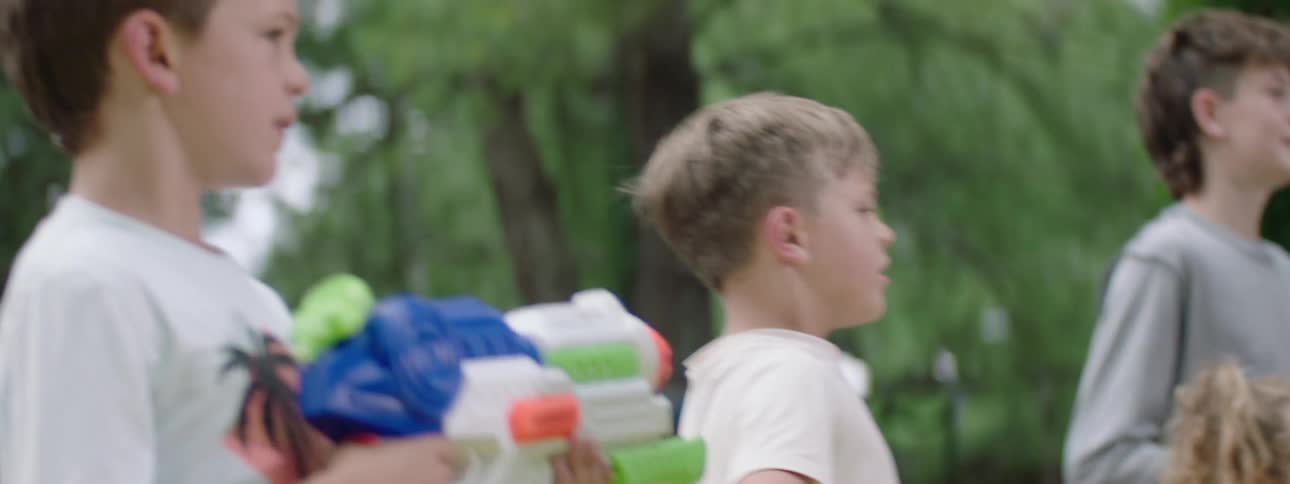 The video captures a group of children outdoors seemingly engaged in a lively activity a boy with a mullet hairstyle stands prominently in the center holding a football while other children around him are also visible some holding water guns