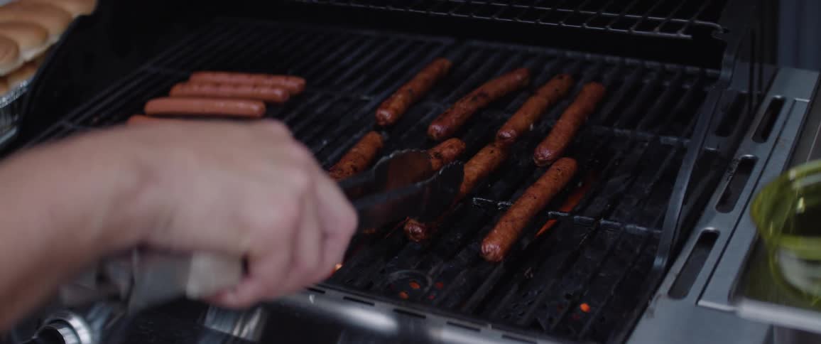 A man grills sausages on an outdoor grill at midday wearing a baseball hat and sunglasses