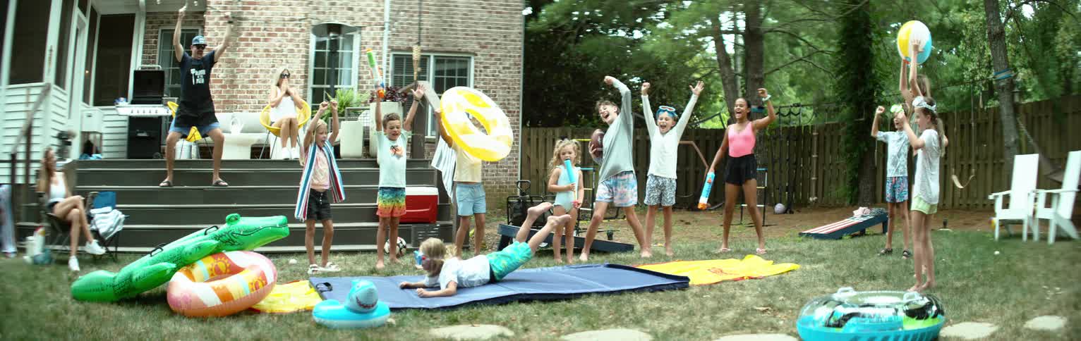 The video displays a vibrant outdoor scene with a group of children and adults in a backyard setting several children are engaged in water play with inflatable toys and a yellow and blue water slide visible on the grass