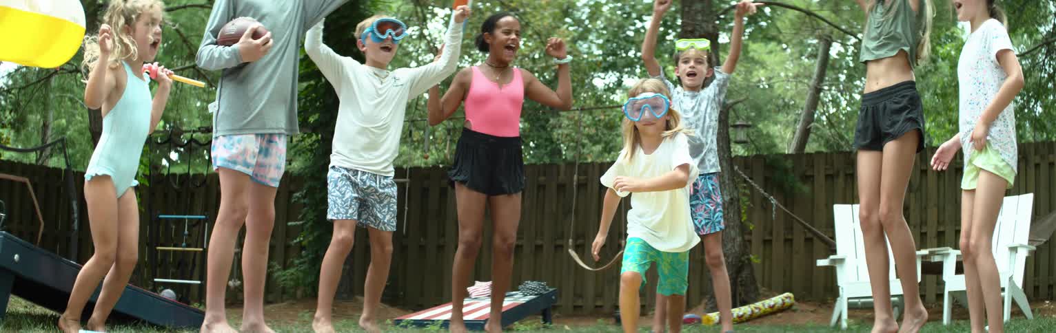 A group of excited children in swimsuits and goggles jump and cheer in a lively backyard setting surrounded by trees and summer play equipment