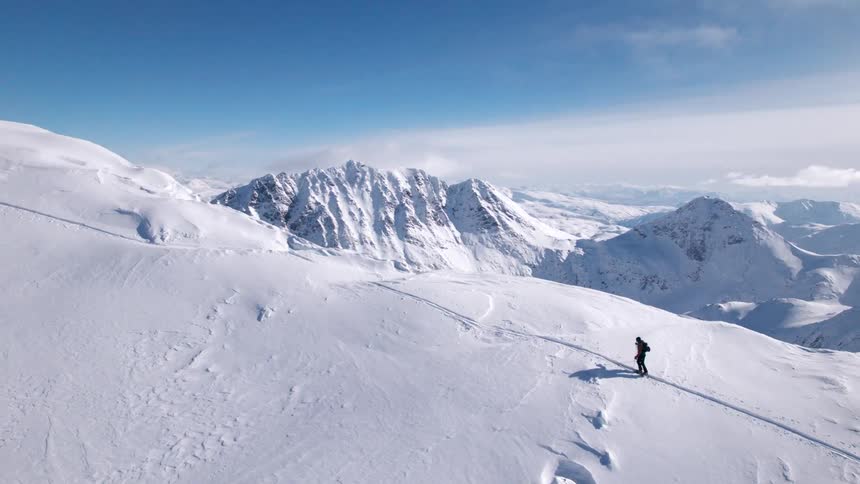 The video shows an individual crosscountry skiing down a snowy mountain slope on a bright clear day the sky is a vibrant blue with sparse clouds complementing the extensive white snow covering the mountains