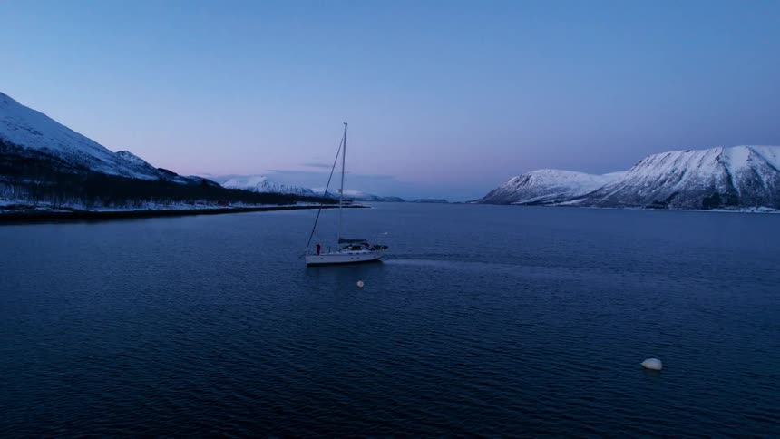 People stand in a sailboat floating off a forested shore after dusk during winter