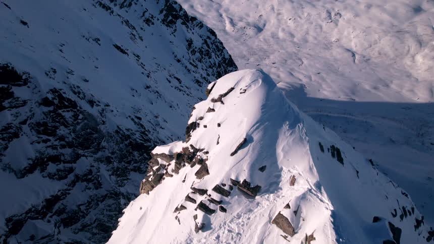 The video presents an extreme closeup highangle aerial view of a snowcovered mountain peak showing jagged rocks protruding through the pristine white snow in the background vast expanses of snowladen terrain and other mountain ranges are visible u