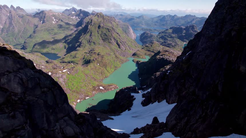 The video features a breathtaking highangle view looking down into a deep mountain pass with snow still clinging to the bottom of the rocky chasm beyond the immediate pass a vibrant turquoise lake stretches out surrounded by steep verdant green moun