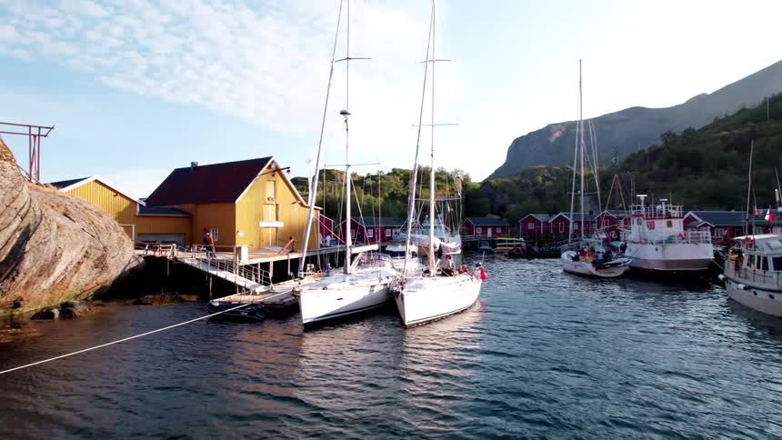 A harbor in a vibrant coastal town is filled with various docked boats including white sailboats and larger vessels surrounded by yellow and red wooden buildings built into rocky cliffs with green mountains and a bright sky in the background