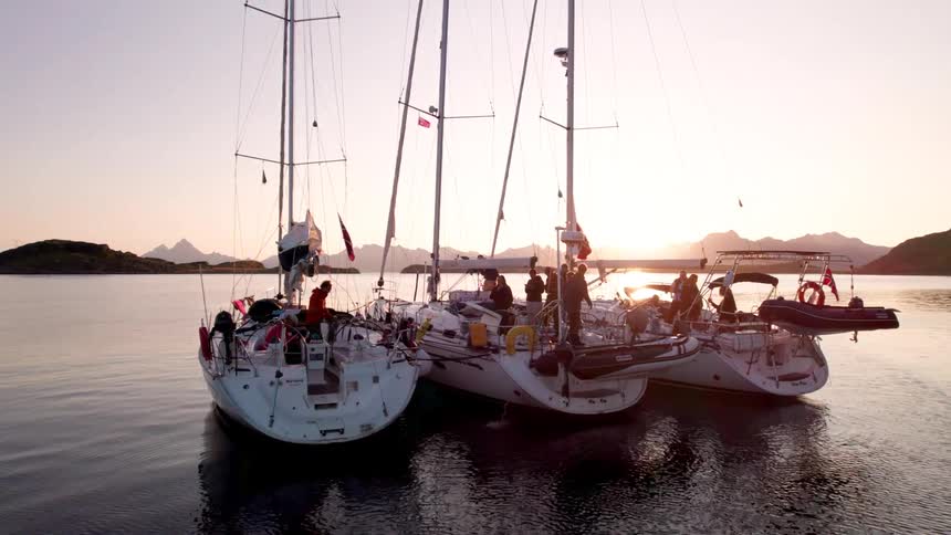 Three sailboats are docked closely side by side near the shore at sunset with people moving about on them