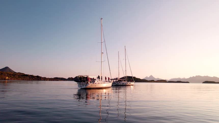 Two sailboats float on calm water with one boat closer to the camera and turning to face away as distant islands and mountains are silhouetted against a bright hazy sky during a sunset