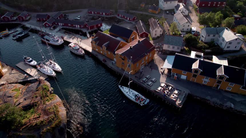 An aerial drone shot captures a charming coastal village nestled among green hills with a marina full of boats and colorful houses illuminated by sunlight