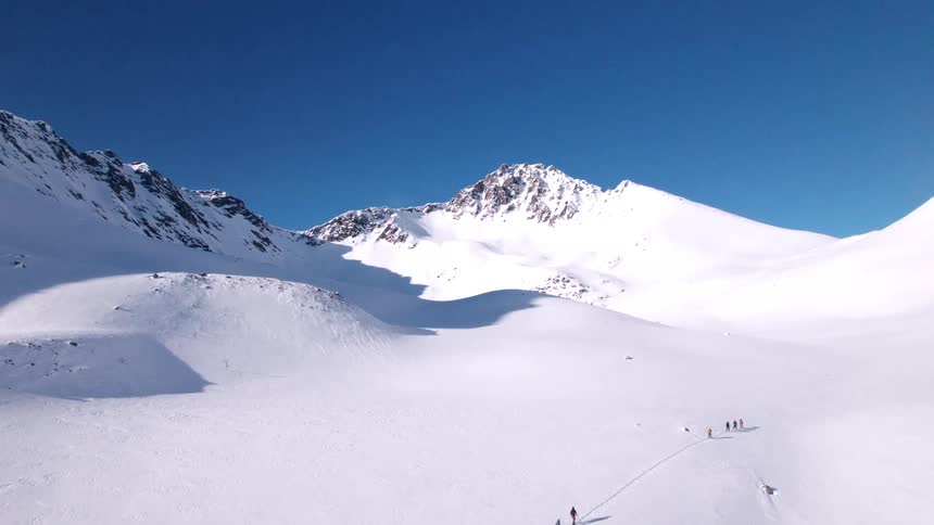 A vast snow covered mountain range stretches under a clear blue sky with a small line of skiers ascending a slope and distant peaks dominating the landscape