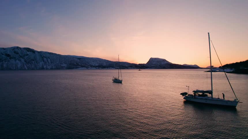 Sailboats drift quietly on calm waters at sunset silhouetted against a glowing horizon with snowlined shores in the distance