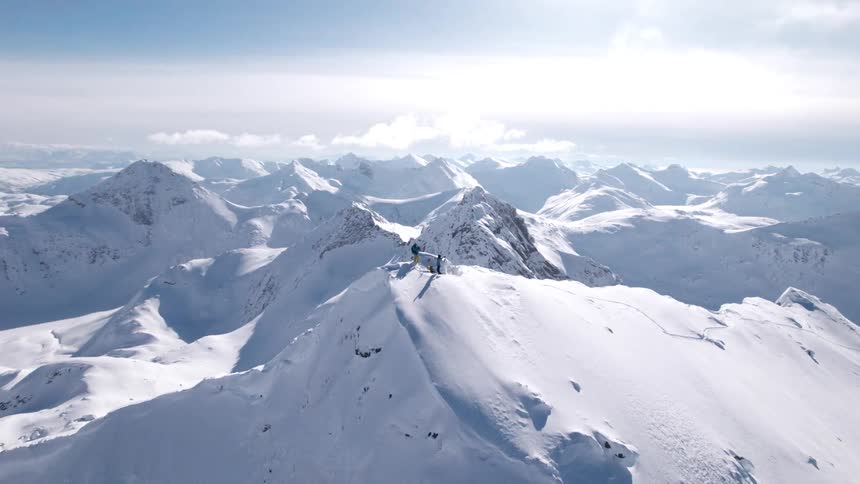 A skier stands atop a pristine snowcovered peak surrounded by a vast panorama of rugged alpine mountains under a clear blue sky