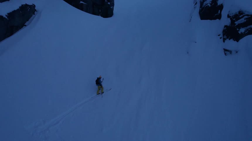 A skier ascends a steep shadowed slope between towering cliffs leaving a solitary trail in the untouched snow