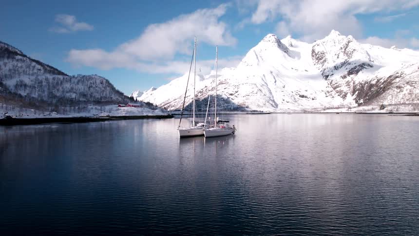 Two sailboats rest peacefully on a glassy fjord surrounded by snowcapped mountains beneath a crisp blue sky