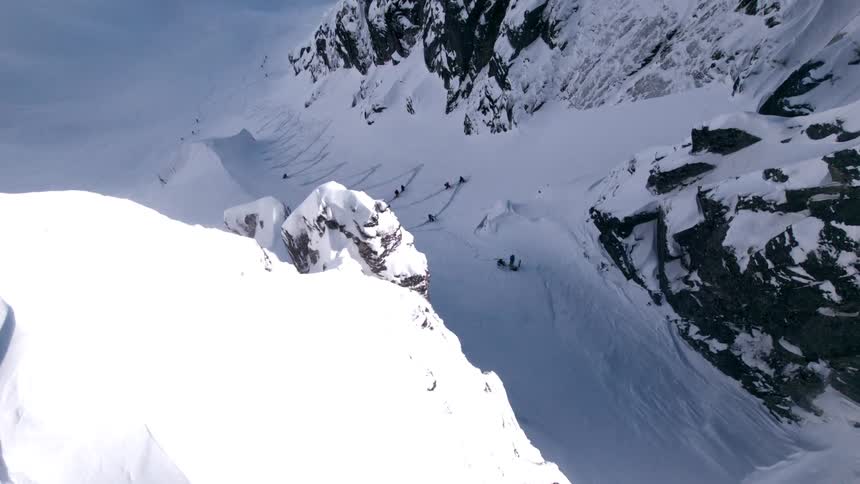 People move along a zigzagging path through the snow in a shadowed hollow on a mountainside on a winter day