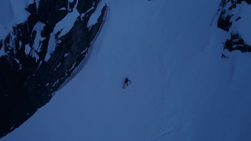 The video shows an extreme highangle shot of a skier making their way down a vast snowcovered slope flanked by towering dark rock faces on either side the scene is dominated by deep blues and whites creating a dramatic and isolated winter mountain