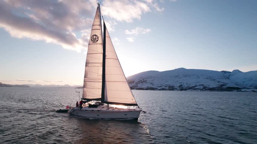A sailboat with white sails glides across a body of water towards the right with snowcapped mountains and a setting sun visible in the background creating a serene and picturesque scene