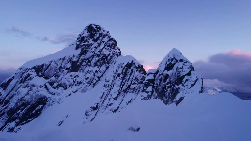 A wide aerial drone shot captures snow covered jagged mountain peaks and vast snowy valleys with faint pink and purple hues in the distant sky