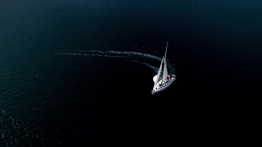An aerial shot captures a sailboat with white sails making a wide turn on dark blue water leaving a prominent wake with snow covered mountains visible in the distant background
