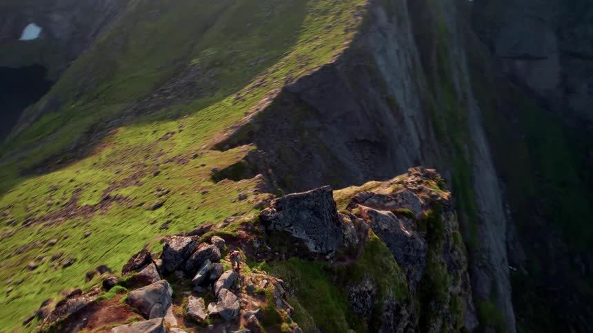 The video provides an aerial perspective of a dramatic mountain ridge steep and rugged covered in a mix of green grass and exposed rock the view also reveals glimpses of a dark body of water and other mountain slopes in the background