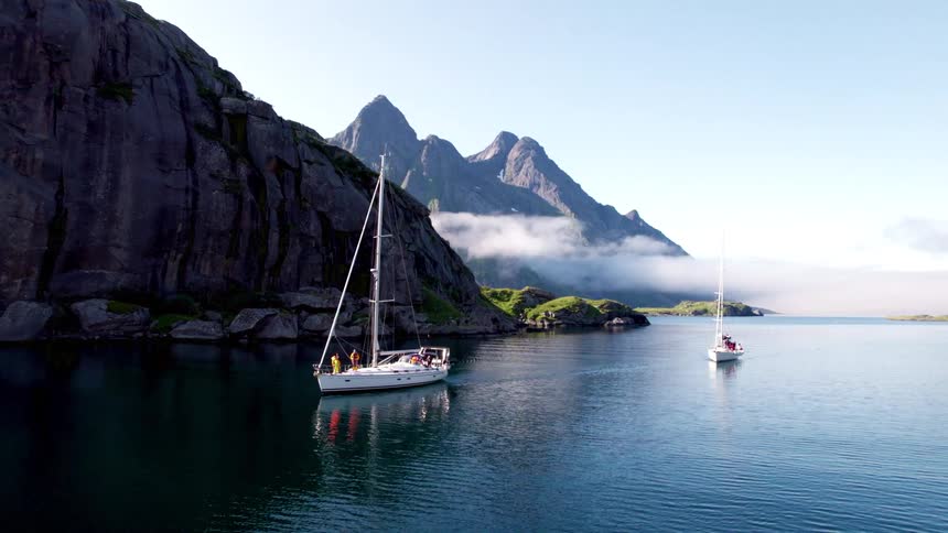 Two sailboats glide across a calm fjord surrounded by towering cliffs and misty mountain peaks