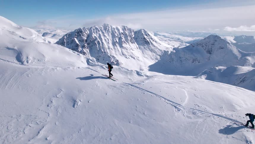The video shows two individuals skiing or snowboarding across a vast snowcovered mountain ridge under a bright sky in the background an extensive range of white jagged peaks stretches into the distance suggesting a remote and expansive winter wonder