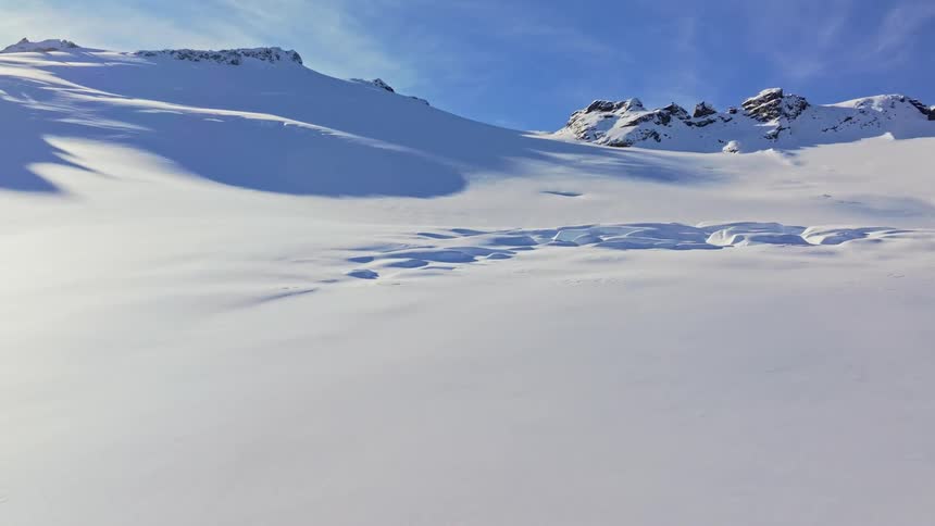 A wide snowy mountain landscape stretches under a bright blue sky with visible tracks and subtle undulations in the snow suggesting a vast untouched wilderness