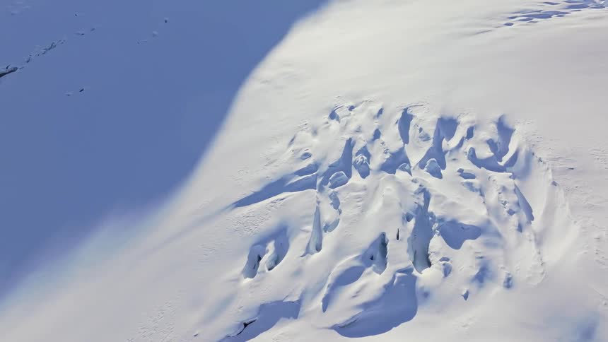 A windswept snowfield reveals jagged ice formations beneath a broad shadow set against a clear blue alpine backdrop