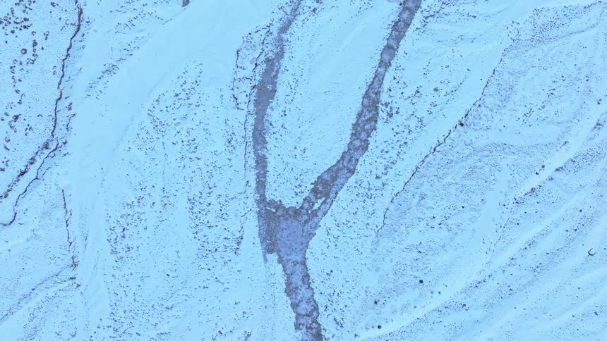 The video displays an abstract highangle view of a textured surface possibly snow or ice with visible lines and cracks a prominent darker crack splits into a y shape at the top contrasting with the predominantly light blue and white hues of the s
