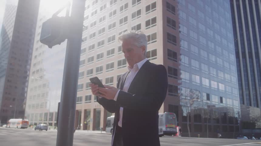A man in a suit stands on a city street corner scrolling on his smartphone