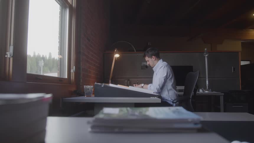 A man works intently at a drafting table in a warmly lit office surrounded by exposed brick walls and natural light streaming through a large window