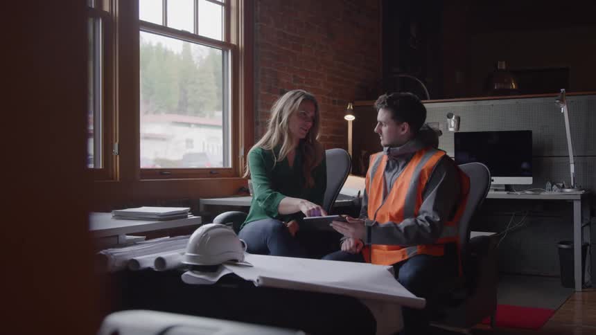 A man in a safety vest sits at a desk in a dimly lit office as a female points to a paper