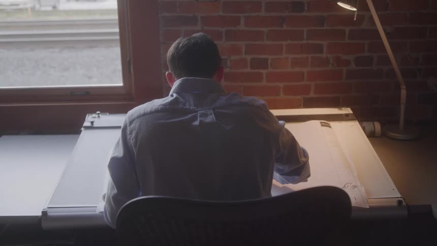 A man sits at a large drafting desk and writes on a schematic
