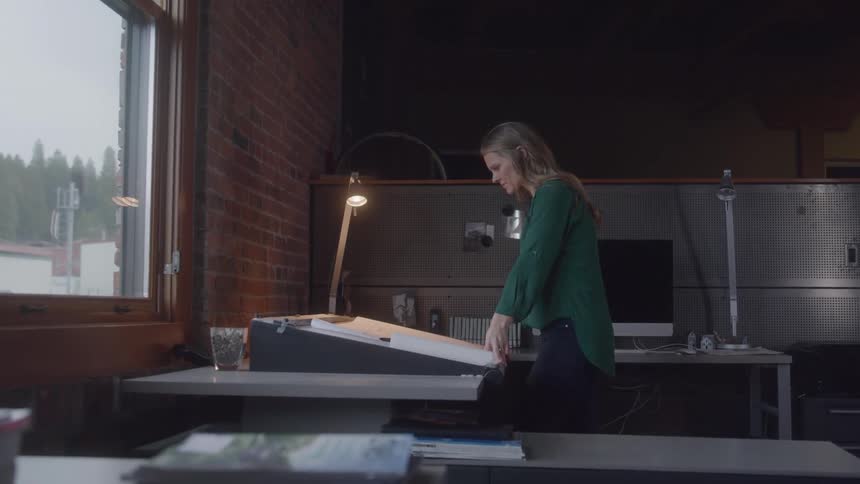 This video captures a side profile of a woman with blonde hair wearing a green blouse and dark pants adjusting a large sheet of paper on an angled drafting table the scene appears to be an office or studio with exposed brick on the left and a desk with