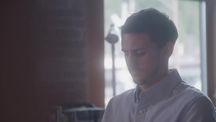 A young adult man with short hair wearing a light colored collared shirt working on an office with a window and a brick wall in the background