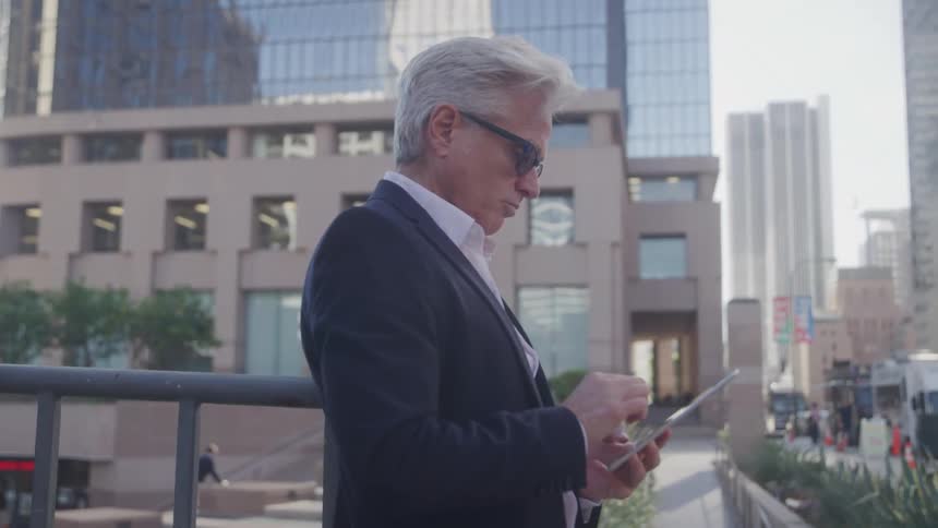 A silverhaired man in business attire stands in a city plaza using a tablet surrounded by tall modern buildings and bustling streets