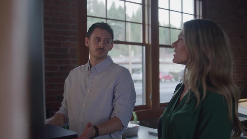 Coworkers engage in a conversation at a desk in an office with brick walls and large windows looking out at a street
