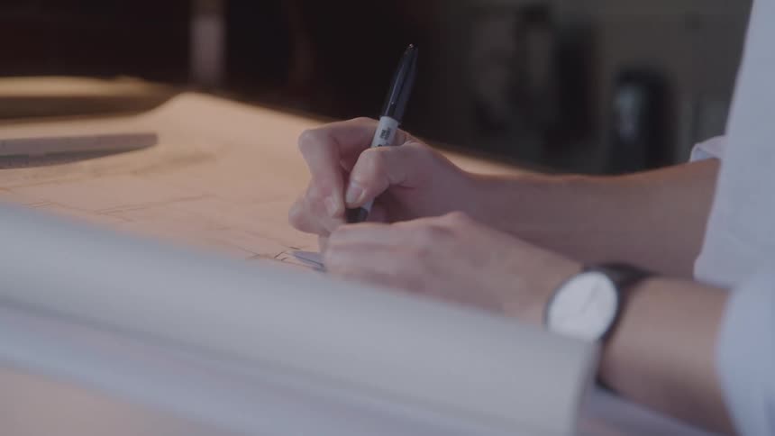 This video offers a closeup of a persons hands one wearing a watch as they meticulously draw or mark on a blueprint or large sheet of paper with a black pen the scene suggests detailed architectural or technical drafting work