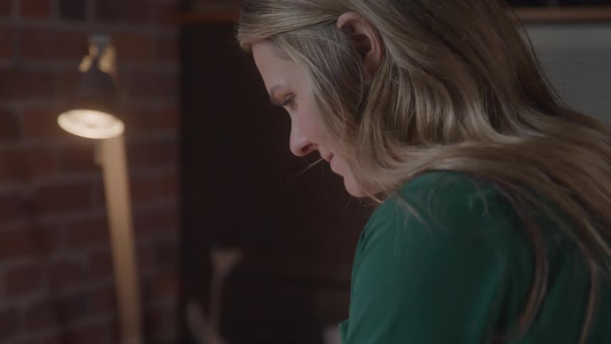 This video features a closeup overtheshoulder view of a woman with long blonde hair wearing a green top intently focused on something below the frame a bright desk lamp is visible to the left illuminating a brick wall in the background suggestin